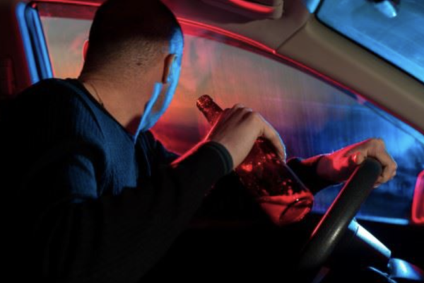 A man in the driver's seat with a beer in his hand looking out the window toward out-of-focus police lights on Blackout Wednesday.